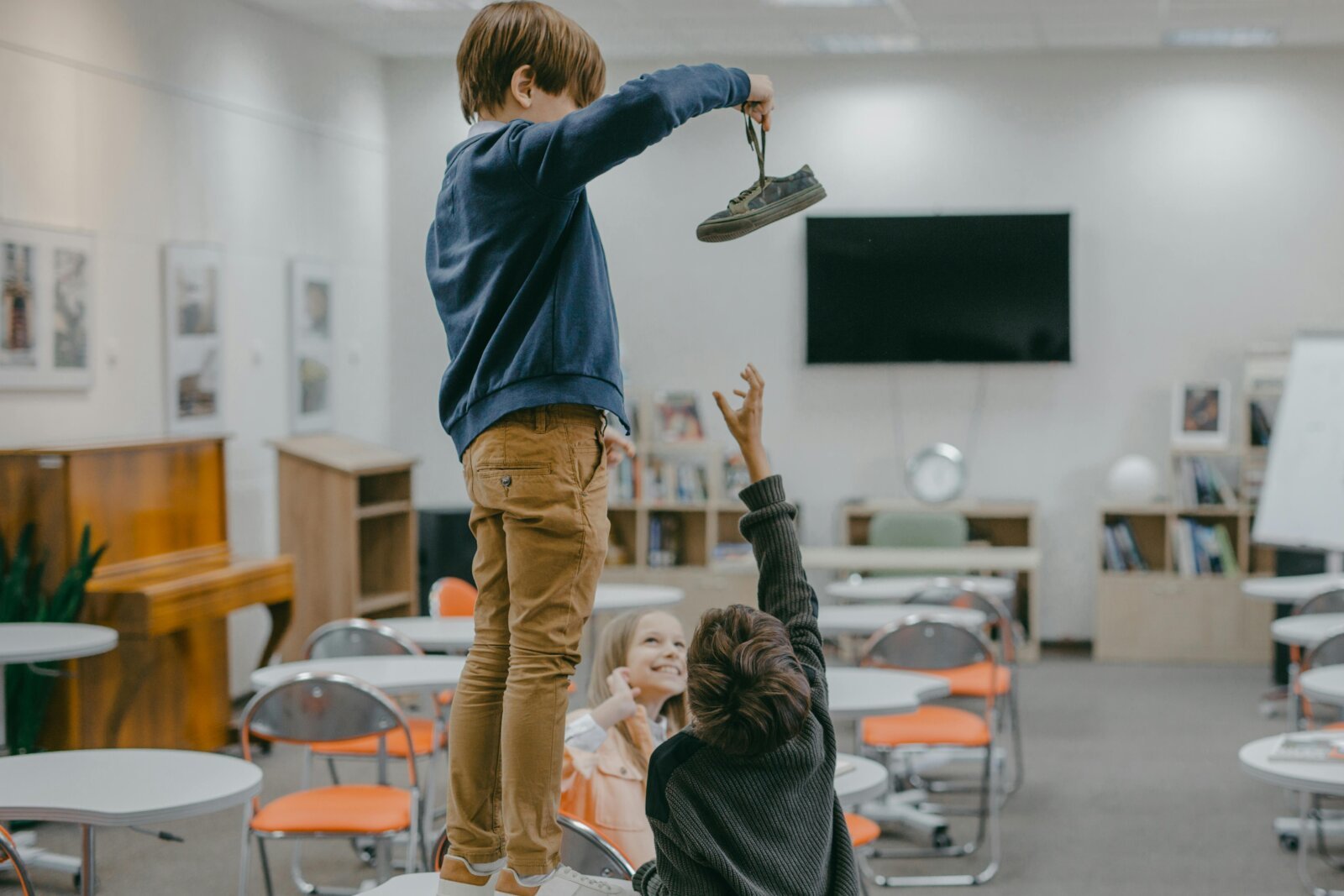Two children stand on a classroom table, one holding a shoe upward while another reaches to help.
