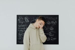 Student in beige hoodie covering face with hand while standing in front of blackboard filled with mathematical equations.