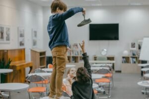 Two children stand on a classroom table, one holding a shoe upward while another reaches to help.