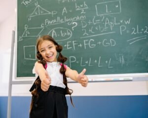 Young girl with braided hair giving thumbs up in classroom with green chalkboard showing math equations.